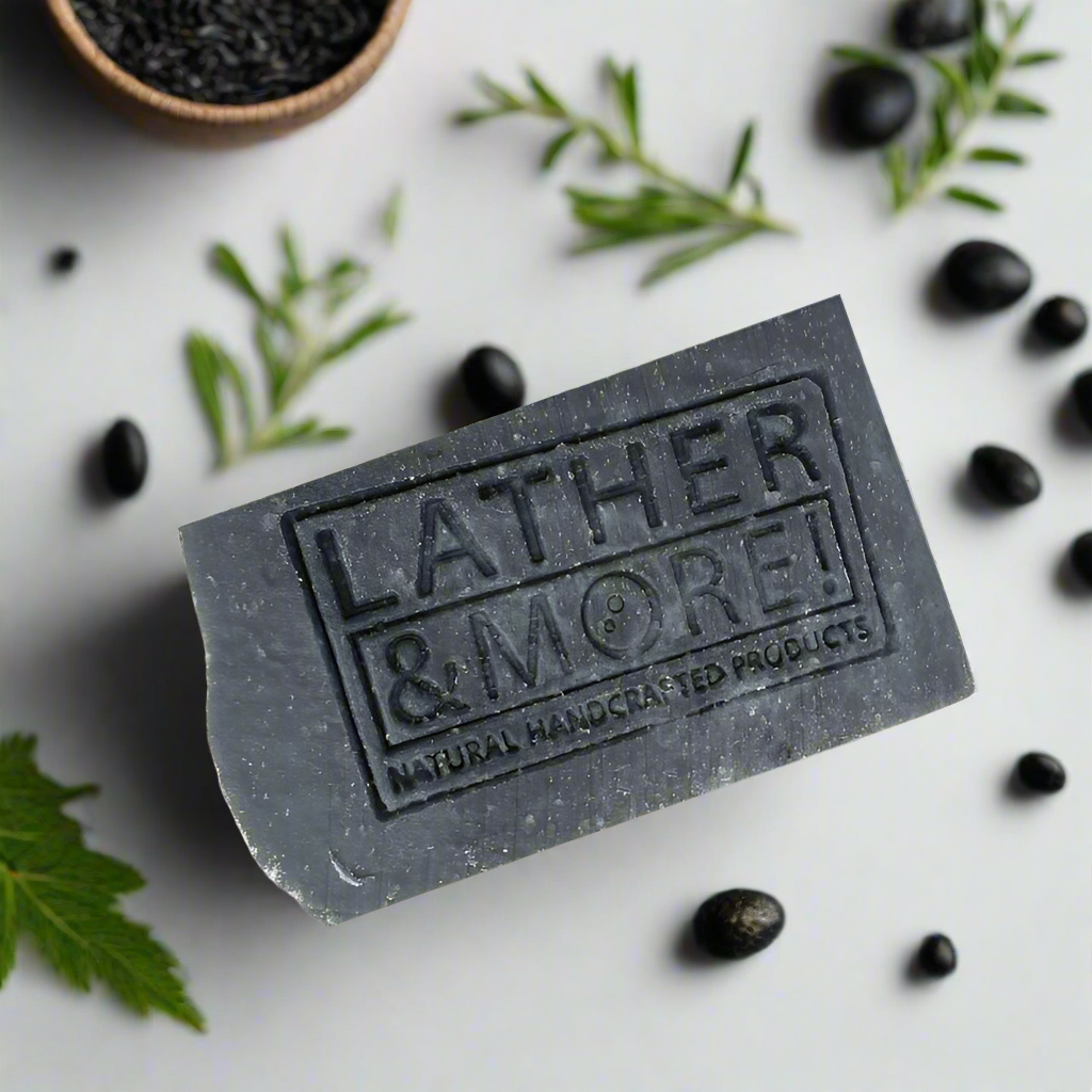 Dark charcoal grey soap bar with company logo stamped on front side of bar. Surrounded by black cumin seeds and fresh greenery.