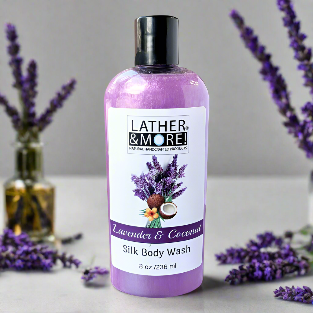 Lavender & Coconut Silk Body Wash - Purple shimmer colored liquid soap in a clear bottle with black pop top cap, Company brand logo with product name and images of lavender flowers and coconuts on front of label on a White  countertop, surrounded by fresh lavender buds on a grey Background