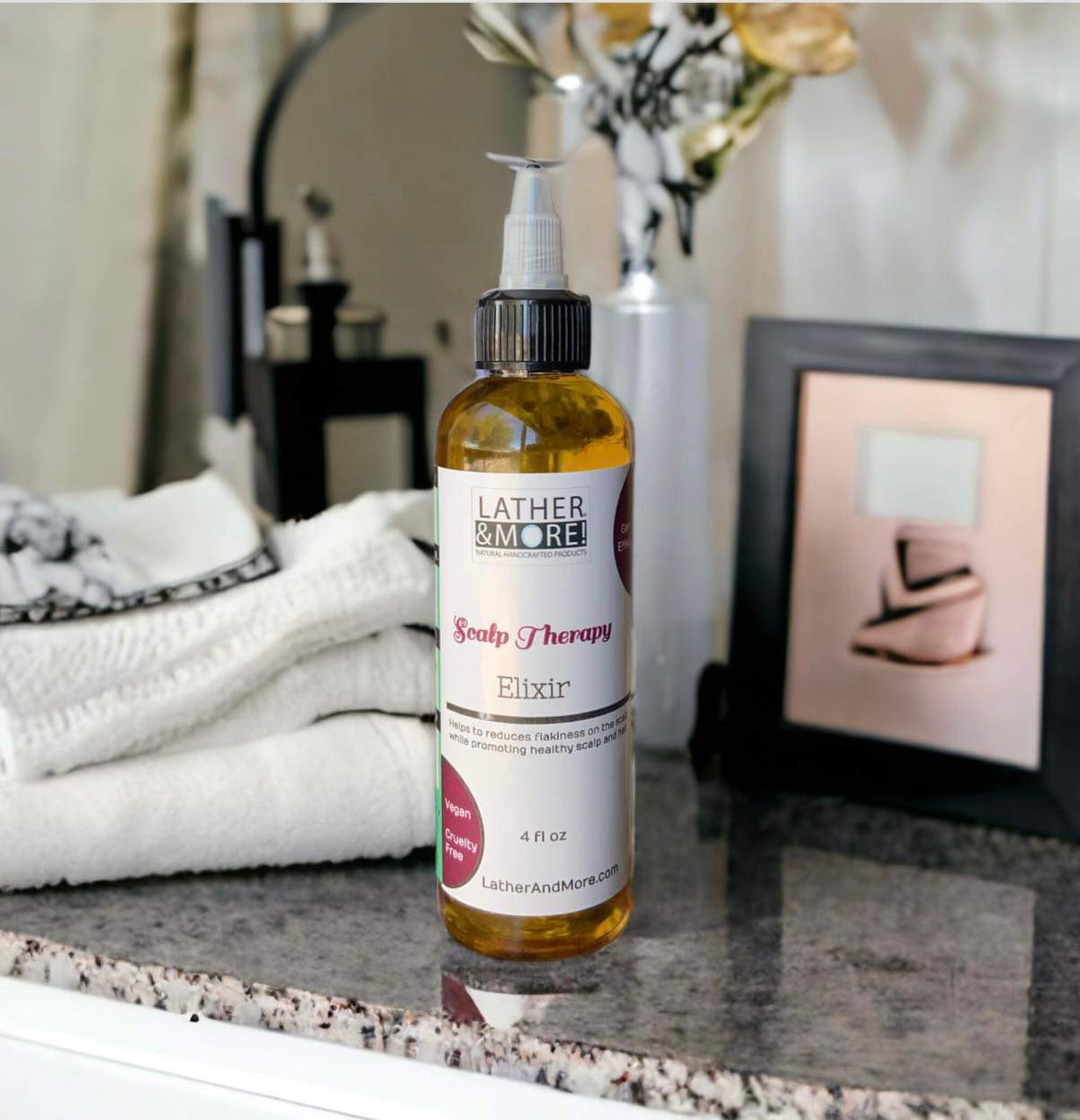 A bottle of Scalp Therapy Elixir by Lather and More placed on a bathroom countertop with a towel and decorative items in the background.