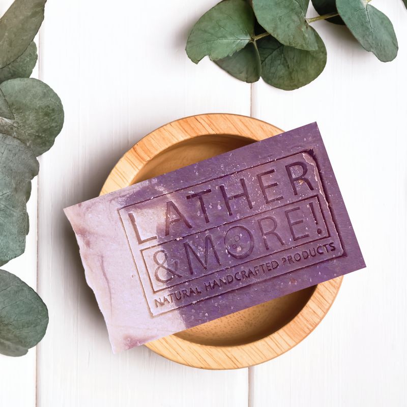 New Money Men's Soap - green top, cream bottom, gold mica line separating the two colors. Soap is sitting in a wooden soap dosh, against a white background with green plant leaves.