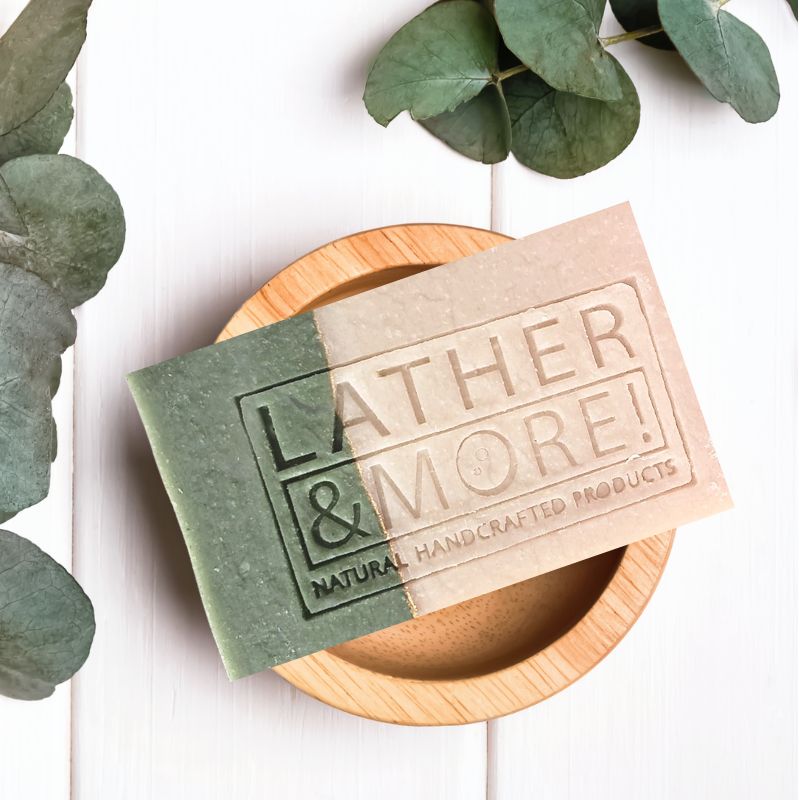A bar of soap with 'LATHER & MORE' engraved on it, placed on a wooden dish with green leaves around it, suggesting natural handcrafted products.