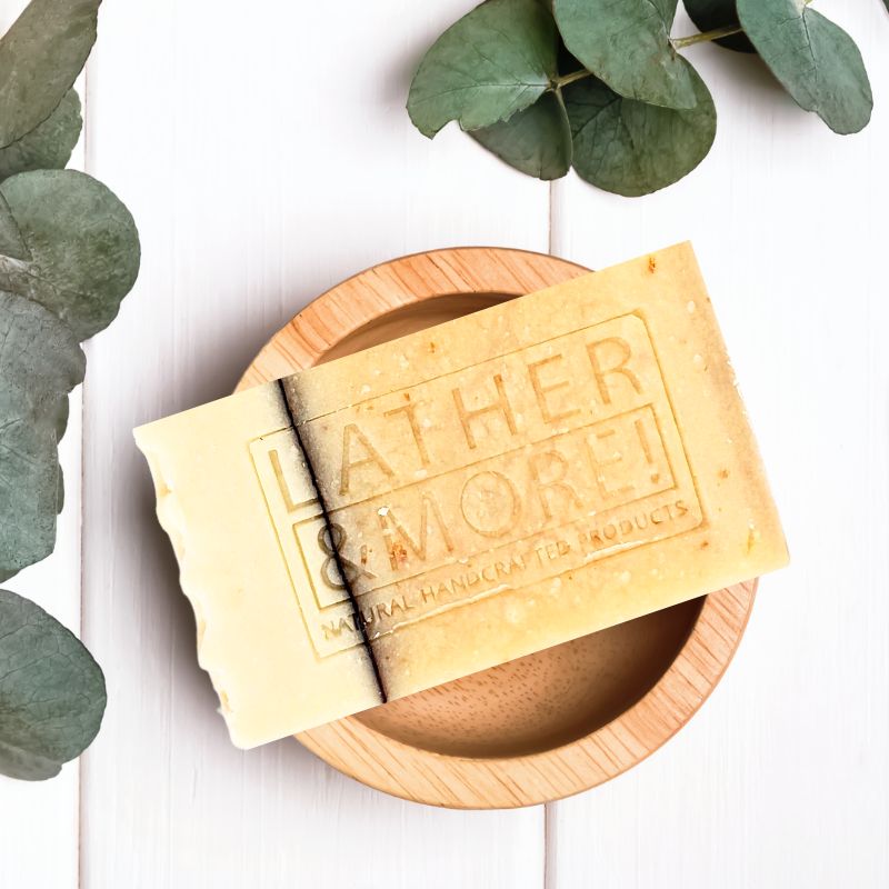 A bar of Oatmeal Milk and Honey Soap resting on a wooden dish, with part of the soap bar lifted to reveal the inside. The soap is light yellow with exfoliating particles visible, and the branding 'LATHER & MORE' is embossed on the top.