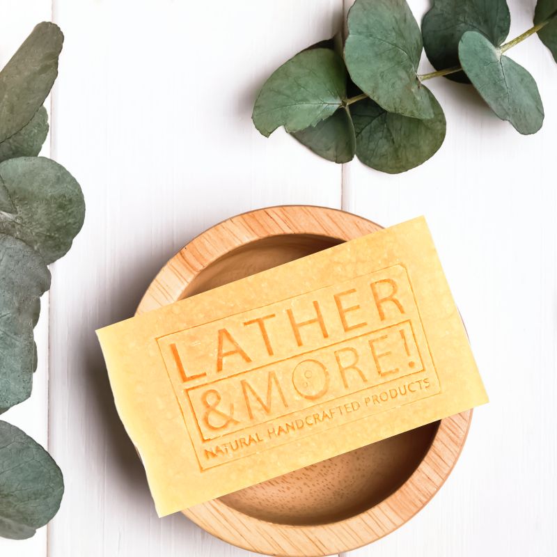 A bar of soap with 'LATHER & MORE' embossed on it, placed on a wooden dish, surrounded by green leaves on a white background.